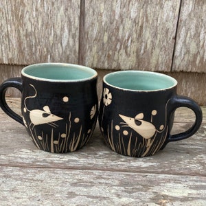 May include: Two black ceramic mugs with a light teal interior. Each mug features a cream-colored mouse illustration among grass and flowers. The mugs have a rounded shape with a handle.