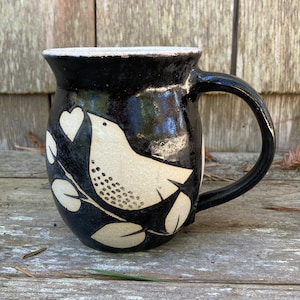 May include: Black ceramic mug with a white bird design. The bird is perched on a branch with leaves and has a heart in its beak.