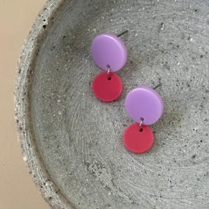 Puede incluir: Un par de pendientes de botón con dos discos circulares superpuestos. El disco superior es de color violeta claro sólido, mientras que el disco inferior, más pequeño, es de color rosa brillante sólido. Incluyen un poste metálico.