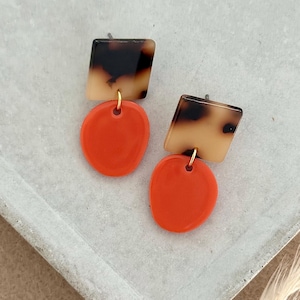 Op de afbeelding: Oorbellen met een vierkante schildpad-top en een ronde oranje onderkant, verbonden door een kleine gouden ring. De oorbellen worden gepresenteerd op een grijze tray.