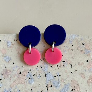 Puede incluir: Un par de pendientes modernos con un disco azul marino conectado a un disco rosa brillante más pequeño. Los pendientes están suspendidos de ganchos plateados. El diseño es minimalista y geométrico, con un toque de color.