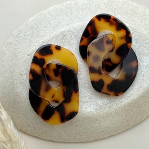 Op de afbeelding: Een paar oorbellen met schildpadpatroon. Elke oorbel heeft een verbonden, abstract ontwerp met amberkleurige en donkerbruine tinten. De oorbellen worden gepresenteerd op een lichtgrijze, getextureerde ondergrond.