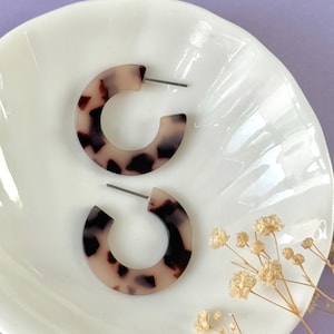 Può includere: Un paio di orecchini a cerchio effetto tartaruga con chiusura a perno. Gli orecchini sono di colore beige chiaro con macchie marrone scuro. Sono esposti su un piatto bianco smerlato su sfondo viola.