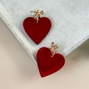 Peut inclure: Une paire de boucles d'oreilles rouges en forme de cœur avec des détails de nœuds dorés. Les boucles d'oreilles sont d'un rouge vif et ont une finition brillante. Les nœuds sont attachés en haut de chaque cœur.