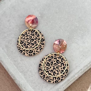 Könnte beinhalten: Ein Paar Ohrhänger mit einem beigefarbenen Kreis mit schwarzen Leopardenflecken, einem goldfarbenen Reifen und einem marmorierten rosa und beigen Oberteil. Die Ohrringe werden auf einer grauen Oberfläche präsentiert.