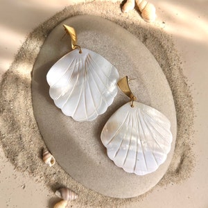 Peut inclure: Une paire de boucles d'oreilles en forme de coquillage dorées et blanches. Les boucles d'oreilles sont en forme de coquillages et ont un crochet en métal doré.
