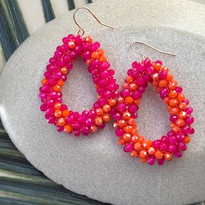 May include: A pair of teardrop-shaped earrings. The earrings are made of small, vibrant pink and orange beads. The beads are arranged to create a textured surface. The ear wires are gold-colored.