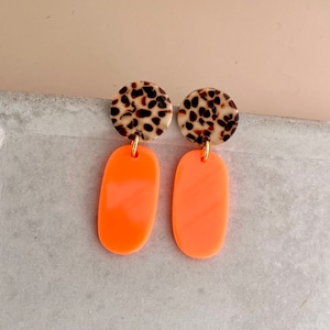 Könnte beinhalten: Ein Paar Ohrringe mit einem kreisförmigen Oberteil in Schildpattmuster und einem leuchtend orangefarbenen, länglichen Unterteil. Die Ohrringe sind mit einer goldfarbenen Schlaufe verbunden. Der Hintergrund ist hellbeige.