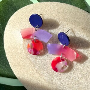 Puede incluir: Un par de pendientes coloridos con un diseño moderno. Cada pendiente presenta un perno azul marino, una forma geométrica rosa y morada, y un dije circular rojo y blanco. Se muestran sobre una superficie beige.