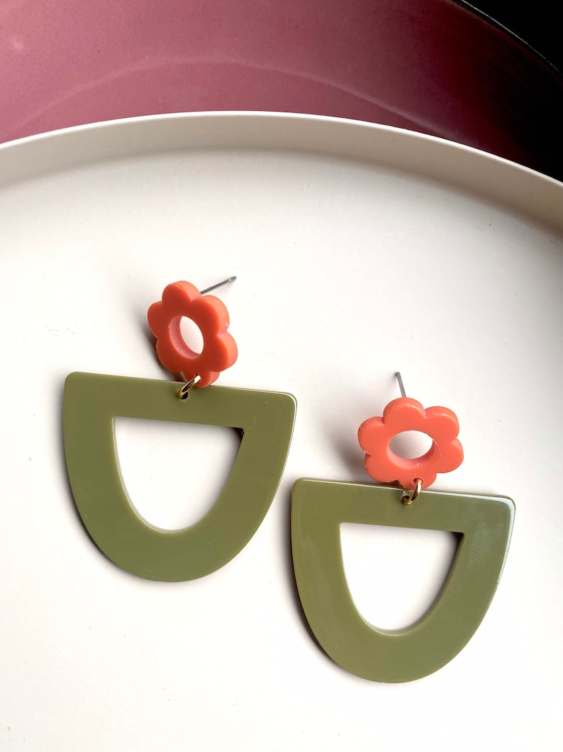 May include: A pair of green and orange statement earrings. The earrings feature a large green arch shape with a smaller circle cut out in the centre. A smaller orange flower charm hangs from the top of the arch.