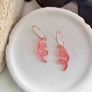 May include: A pair of pink acrylic hoop earrings with a swirl design. The earrings are on a white surface.