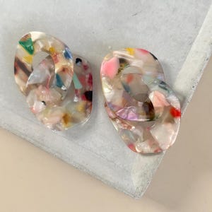 Può includere: Due paia di orecchini ovali collegati. Gli orecchini sono realizzati in un materiale traslucido con macchie colorate di rosa, verde, giallo e blu. Gli orecchini sono appoggiati su una superficie grigio chiaro.