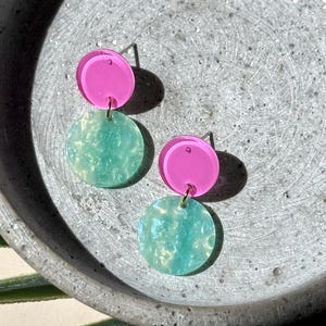 Op de afbeelding: Een paar hangende oorbellen met een kleine, effen felroze ronde knop verbonden aan een grotere, doorschijnende mintgroene ronde hanger met een textuurrijke, gemarmerde afwerking, bevestigd met metalen stiftjes en ringetjes.