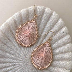 May include: A pair of pink and gold teardrop-shaped earrings. The earrings are made of wire wrapped with pink thread and have a gold wire frame.