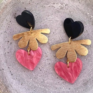 Könnte beinhalten: Ein Paar goldene und rosafarbene Ohrringe in Herzform mit schwarzen Herz-Akzenten. Die Ohrringe haben einen goldenen, blumenförmigen Anhänger und ein rosafarbenes Herz mit Marmorierung.