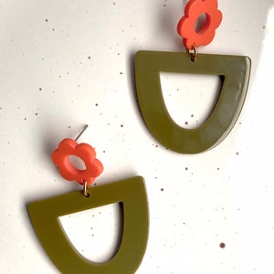 May include: A pair of olive green and orange earrings. Each earring features a half-circle shape with a cut-out in the centre and a small orange flower charm.
