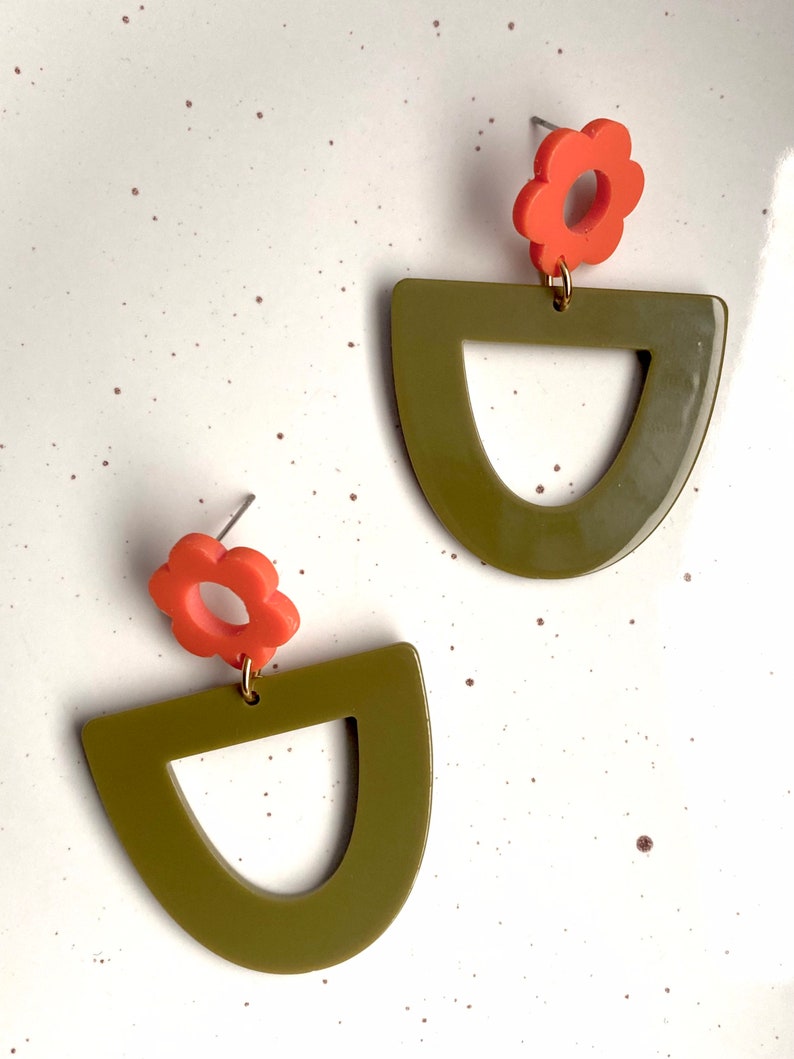 May include: A pair of olive green and orange earrings. Each earring features a half-circle shape with a cut-out in the centre and a small orange flower charm.