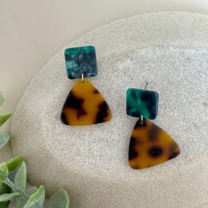 Puede incluir: Un par de pendientes geométricos. Cada pendiente presenta una parte superior cuadrada de color verde oscuro y un colgante triangular de carey. Los pendientes se exhiben sobre una superficie de piedra gris claro, con follaje verde en el fondo.