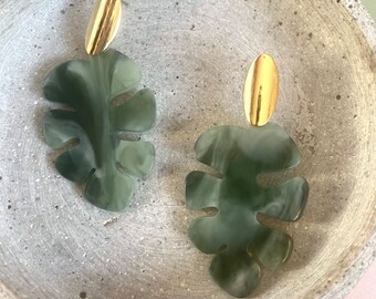 Pode incluir: Um par de brincos de folha verde e dourada. Os brincos são feitos de um material verde marmorizado e têm um acento oval dourado na parte superior.