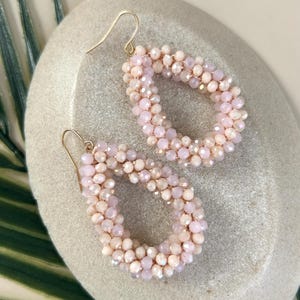 Può includere: Un paio di orecchini pendenti con perline rosa e ganci dorati. Le perline sono piccole e iridescenti, creando un effetto scintillante.