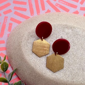 Könnte beinhalten: Ohrringe mit einem bordeauxroten Kreis und einem goldfarbenen, sechseckigen Anhänger. Die Ohrringe werden auf einer hellgrauen, strukturierten Oberfläche mit einem rosa-orangenen Hintergrund präsentiert.