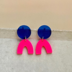 Könnte beinhalten: Ein Paar leuchtende Ohrringe mit einem kreisförmigen blauen Oberteil und einer neonpinkfarbenen Bogenform. Die Ohrringe sind an einem kleinen silbernen Haken befestigt. Das Design ist modern und geometrisch.