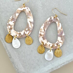 Puede incluir: Pendientes colgantes con un diseño veteado en tonos beige, blanco y lavanda. Cada pendiente tiene un gancho dorado y tres colgantes: dos discos dorados y una lágrima nacarada. Un accesorio elegante.