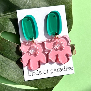 Peut inclure: Boucles d'oreilles composées d'une partie supérieure ovale verte et d'une fleur rose. Les boucles d'oreilles sont reliées par des attaches dorées. Le texte "birds of paradise" est imprimé sur une carte blanche.