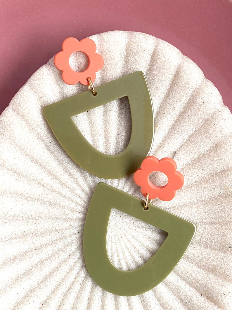 May include: A pair of green and coral earrings. The earrings are shaped like a half-circle with a smaller circle cut out of the centre. The top of each earring is adorned with a coral flower.