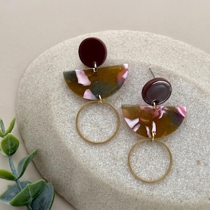 Può includere: Un paio di orecchini fatti a mano con un perno bordeaux scuro, un ciondolo a semicerchio con marmorizzazione verde oliva, rosa e bianca e un cerchio dorato. Gli orecchini sono esposti su una pietra grigio chiaro.