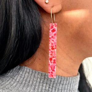 Op de afbeelding: Een gouden oorring met een lange, rechthoekige roze en rode bedel met een patroon. De bedel heeft een gespikkeld ontwerp. De oorring hangt aan een oor. De bedel is ongeveer 5 cm lang.