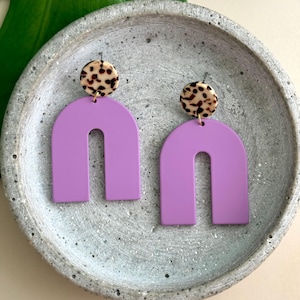 Puede incluir: Un par de pendientes colgantes que presentan un poste circular con un patrón de leopardo marrón y negro unido a un colgante en forma de arco de color morado lavanda sólido. Los pendientes tienen un acabado mate suave en las piezas moradas.