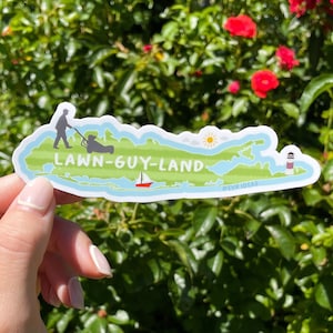 Op de afbeelding: Een sticker in de vorm van Long Island, met de tekst "Lawn-Guy-Land". De sticker toont illustraties van een persoon die een gazon maait, een zeilboot, een vuurtoren en een zon. De sticker wordt vastgehouden tegen een achtergrond van groene bladeren en rode bloemen.