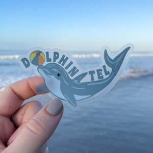 May include: A clear sticker with a blue dolphin and the text "Dolphin Tel" with a yellow and orange beach ball in the text. The sticker is being held in a hand against a blue sky and ocean background.