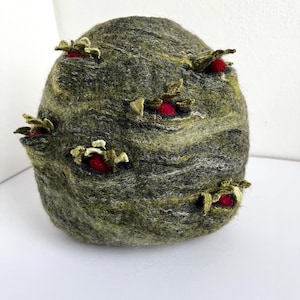 Può includere: Una scultura rotonda in feltro con una superficie testurizzata verde e grigia. La scultura ha cinque fiori in feltro rosso con steli in feltro verde che sporgono dalla superficie.