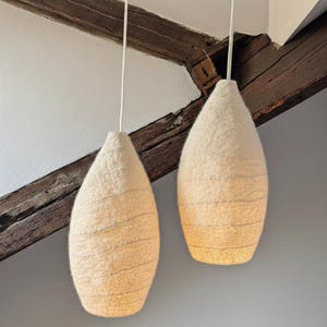 Könnte beinhalten: Zwei cremefarbene Pendelleuchten in Tropfenform aus einem strukturierten Material mit horizontalen Linien. Sie hängen an weißen Kabeln. Der Hintergrund zeigt Holzbalken und eine weiße Decke.