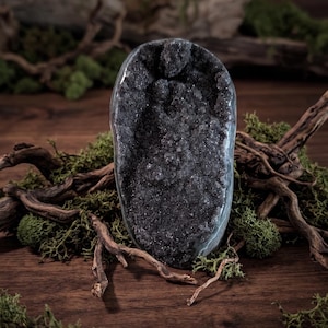 Könnte beinhalten: Eine dunkel anthrazitfarbene Geode mit einem hohlen Zentrum, das mit kleinen, funkelnden kristallinen Strukturen gefüllt ist. Die Außenseite des Steins ist glatt und abgerundet und bildet einen Kontrast zum rauen, texturierten und glitzernden Innenraum.
