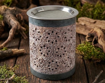 May include: A cylindrical, carved stone tea light holder with a stainless steel rim and mesh screen. The holder features a repeating floral pattern and is set on a wooden surface with moss and branches.