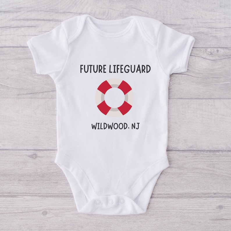 Peut inclure: Combinaison blanche pour b&eacute;b&eacute; avec le texte "FUTURE LIFEGUARD" et un dessin de bou&eacute;e de sauvetage rouge et blanche. Le texte "WILDWOOD. NJ" est imprim&eacute; sous le dessin.