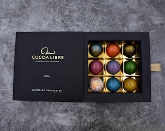 Puede incluir: Una caja negra con un logotipo dorado que dice "Cocoa Libre Allergy Friendly Chocolate" y "Handmade Chocolates". La caja contiene 12 chocolates surtidos en un patrón de cuadrícula.