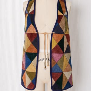 May include: A sleeveless vest constructed from multi-colored suede triangular patches in shades of blue, red, yellow, green, and brown. The garment features a dark blue trim along the edges and a thin tan leather tie closure at the center front.