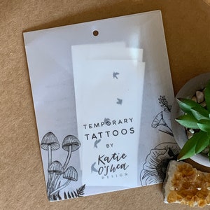 May include: A white package with a hole at the top, containing temporary tattoos with a design of flying birds. The package has a black and white illustration of mushrooms and flowers on the bottom. The text on the package reads "Temporary Tattoos by Katie O'Shea Design."