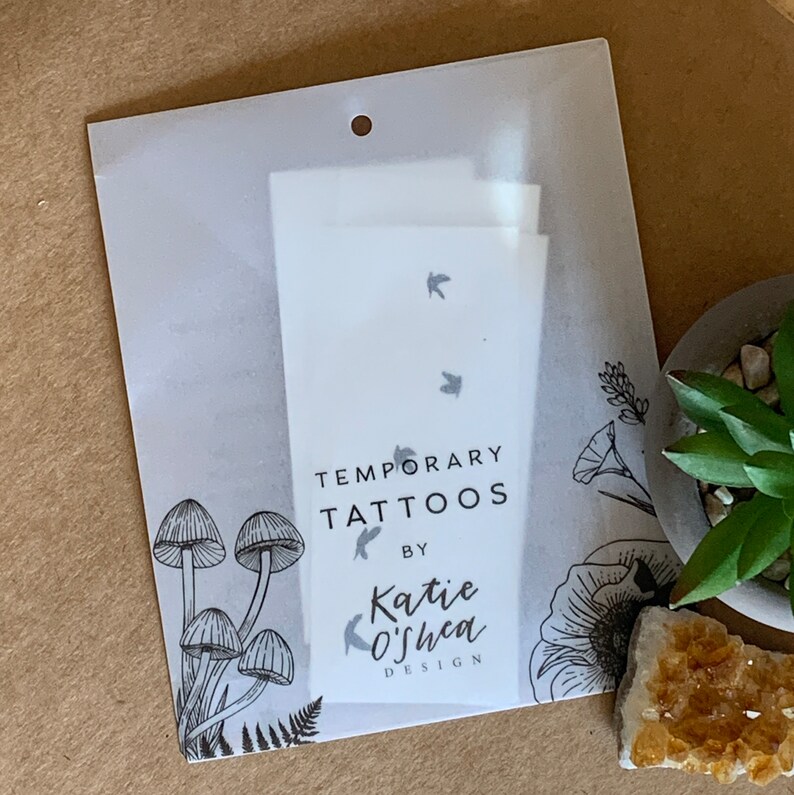 May include: A white package with a hole at the top, containing temporary tattoos with a design of flying birds. The package has a black and white illustration of mushrooms and flowers on the bottom. The text on the package reads "Temporary Tattoos by Katie O'Shea Design."