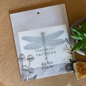 May include: A clear plastic package containing a sheet of temporary tattoos featuring a dragonfly, mushrooms, and flowers. The package has the text "Temporary Tattoos by Katie O'Shea Design".