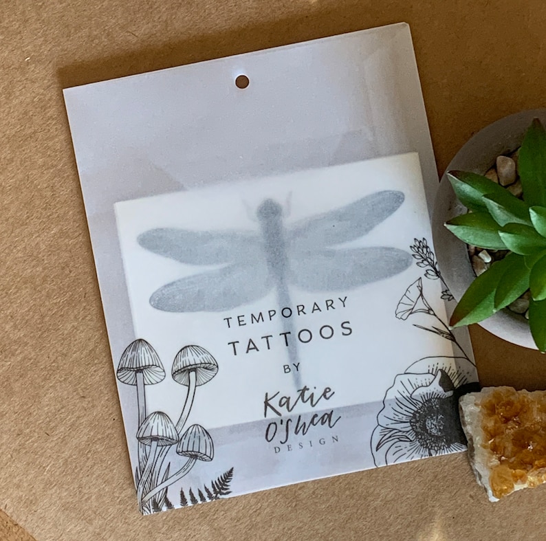 May include: A clear plastic package containing a sheet of temporary tattoos featuring a dragonfly, mushrooms, and flowers. The package has the text "Temporary Tattoos by Katie O'Shea Design".