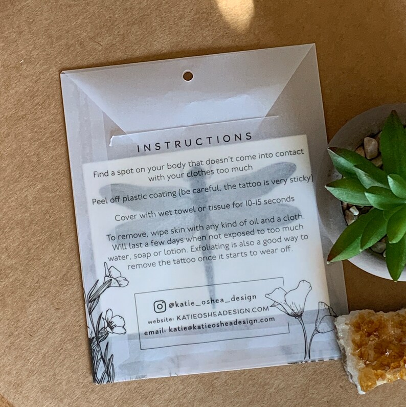 May include: Instructions for applying a temporary tattoo. The instructions are printed on a white card with a black dragonfly and floral design. The text includes information about how to apply and remove the tattoo. The card also includes the social media handle, website, and email address for the company.