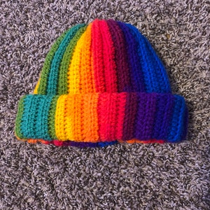 Könnte beinhalten: Eine gestrickte Mütze mit Regenbogenstreifen. Die Mütze ist in verschiedenen Farben gestrickt, darunter Rot, Orange, Gelb, Grün, Blau und Lila.