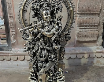 Puede incluir: Una estatua de metal en tono latón de una deidad tocando la flauta, de pie en una postura con las piernas cruzadas. La figura está adornada con joyas intrincadas y una corona, enmarcada por un gran halo circular ornamentado con detalles.