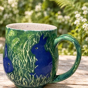 May include: A ceramic mug with a glossy green glaze featuring a hand-painted scene of two dark blue rabbits sitting in tall, light-colored grass. The interior of the mug is a light cream color with small, multi-colored speckles throughout.
