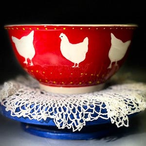 May include: A red ceramic mixing bowl with white chicken silhouettes and yellow polka dots. The bowl sits on a white lace doily and a blue stand. The bowl is a kitchenware item.